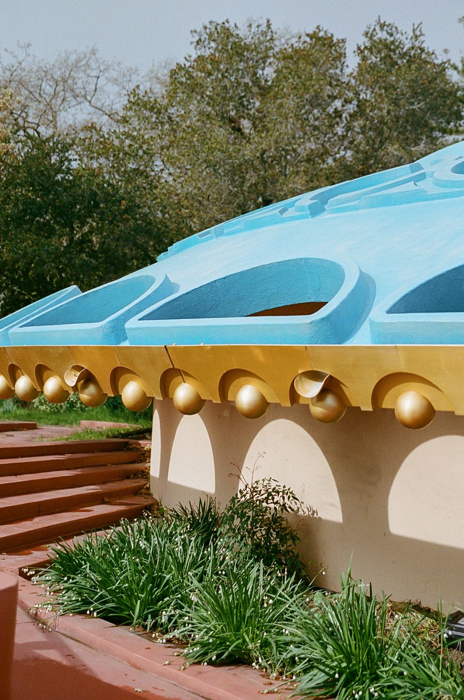 Architectural detail showing a curved blue roof with golden bells and white arches above landscaped steps and greenery.
