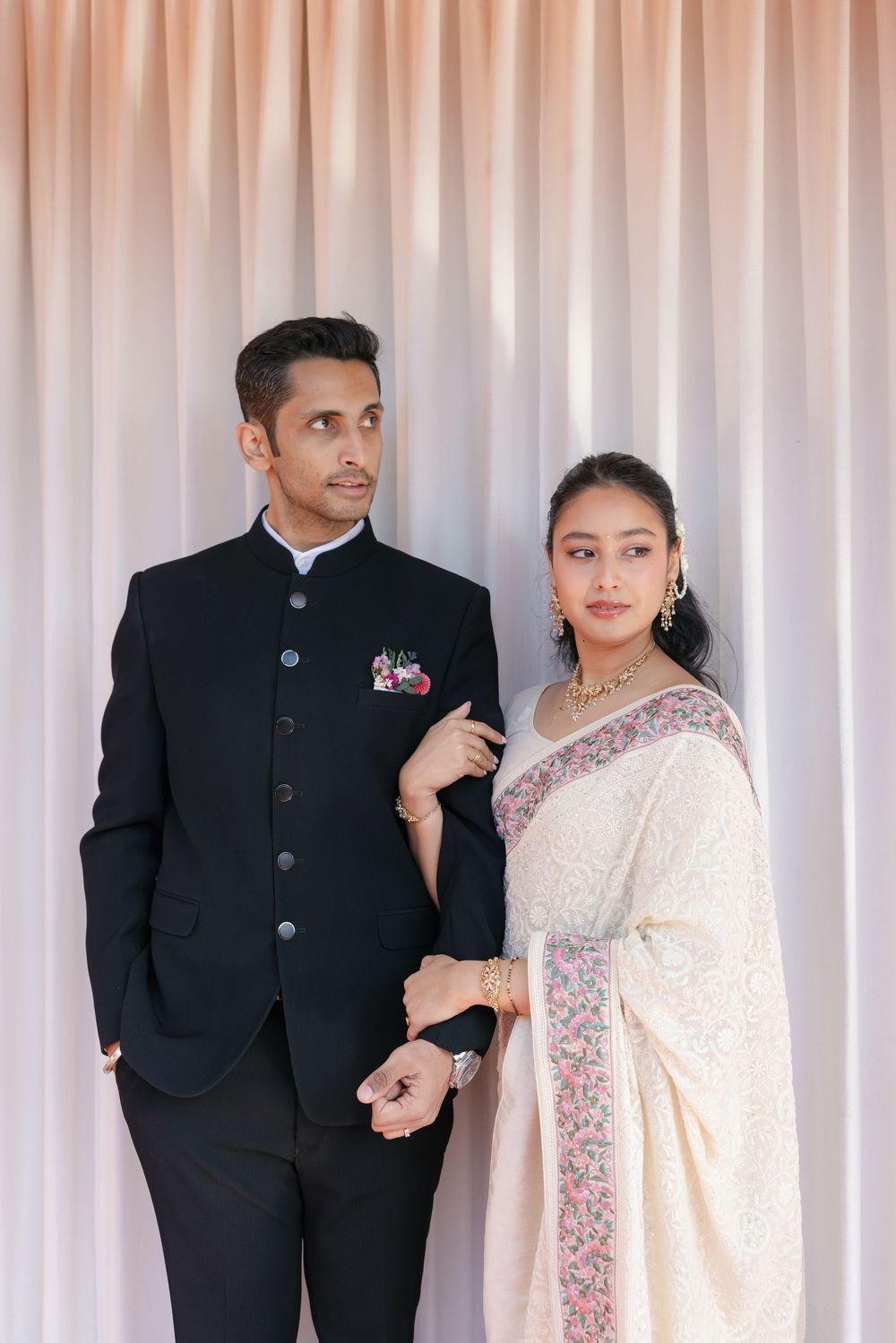 A couple in formal attire, with the man wearing a black suit and the woman in an elegant white sari with embroidered details.
