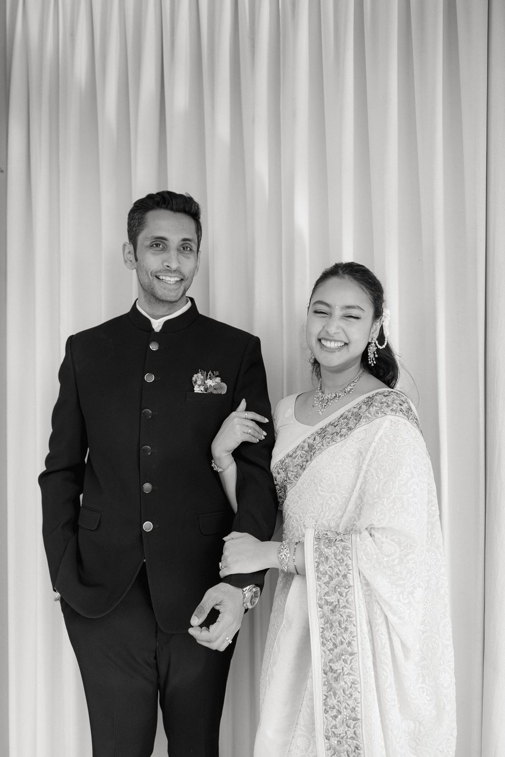 A couple poses in formal Indian attire, with the groom in a black bandhgala and bride in an elegant white saree.