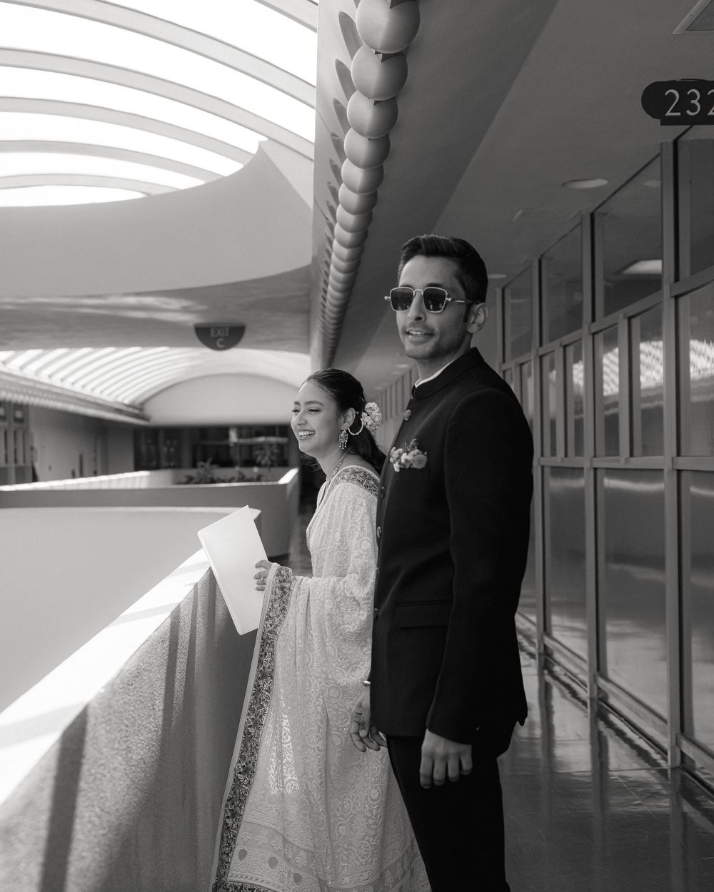 A couple in formal wedding attire poses in a modern hallway with curved architectural details and dramatic lighting.