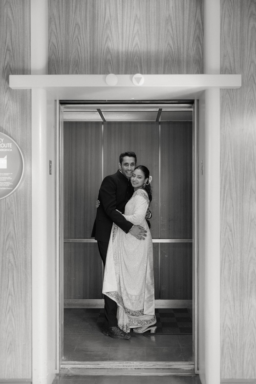 A couple embraces in a vintage-style elevator with wood paneled walls and classic interior details.