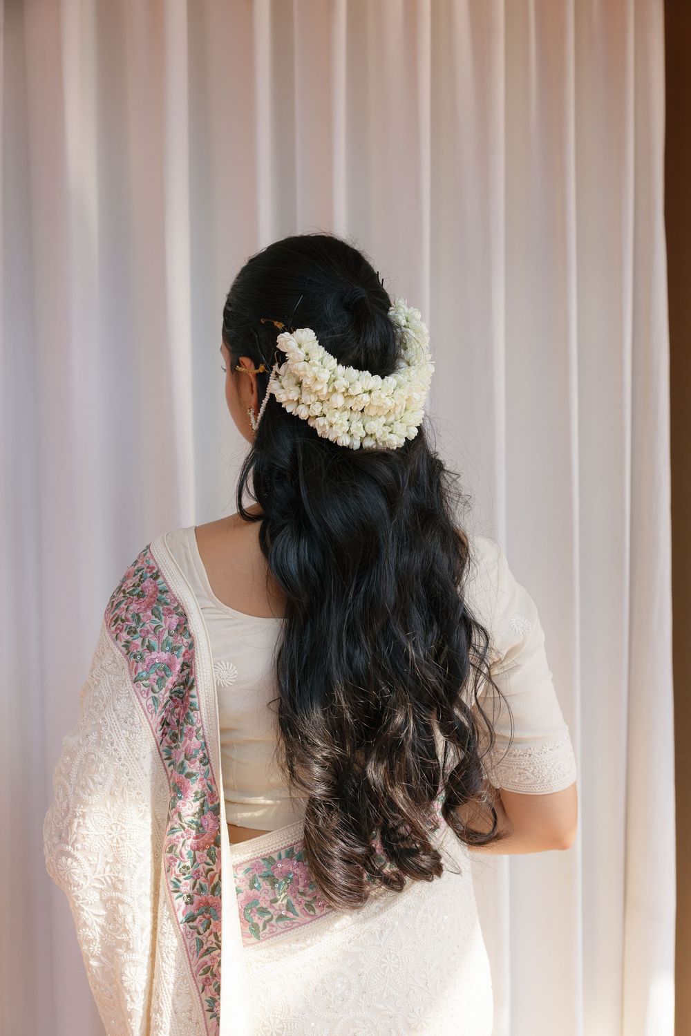 Back view of a woman wearing a white sari with floral details and jasmine flowers in her hair.