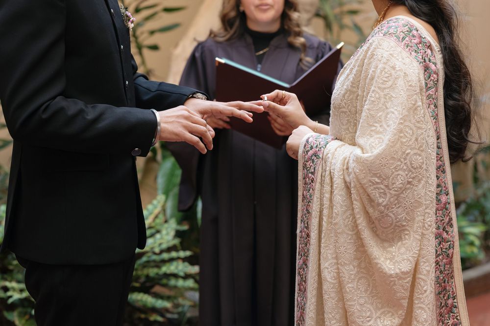 Close-up of hands exchanging wedding rings during an outdoor ceremony with lace dress details.