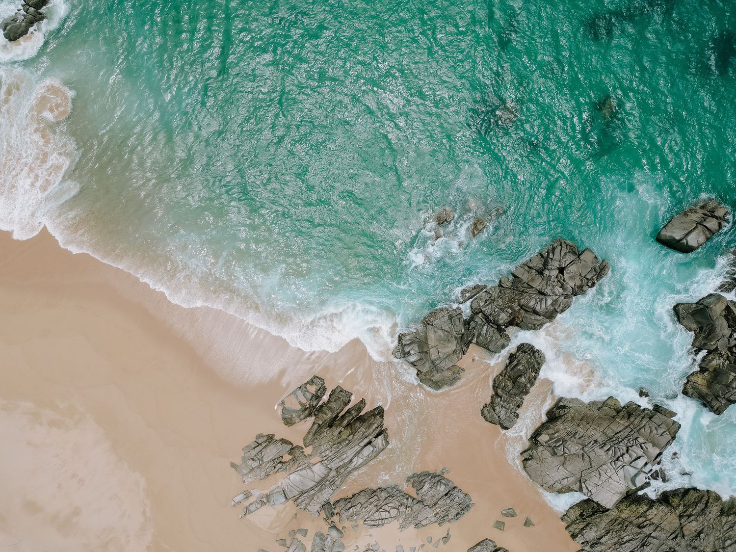 Aerial view of turquoise ocean waves crashing against rocky coastline with sandy beach and white surf.