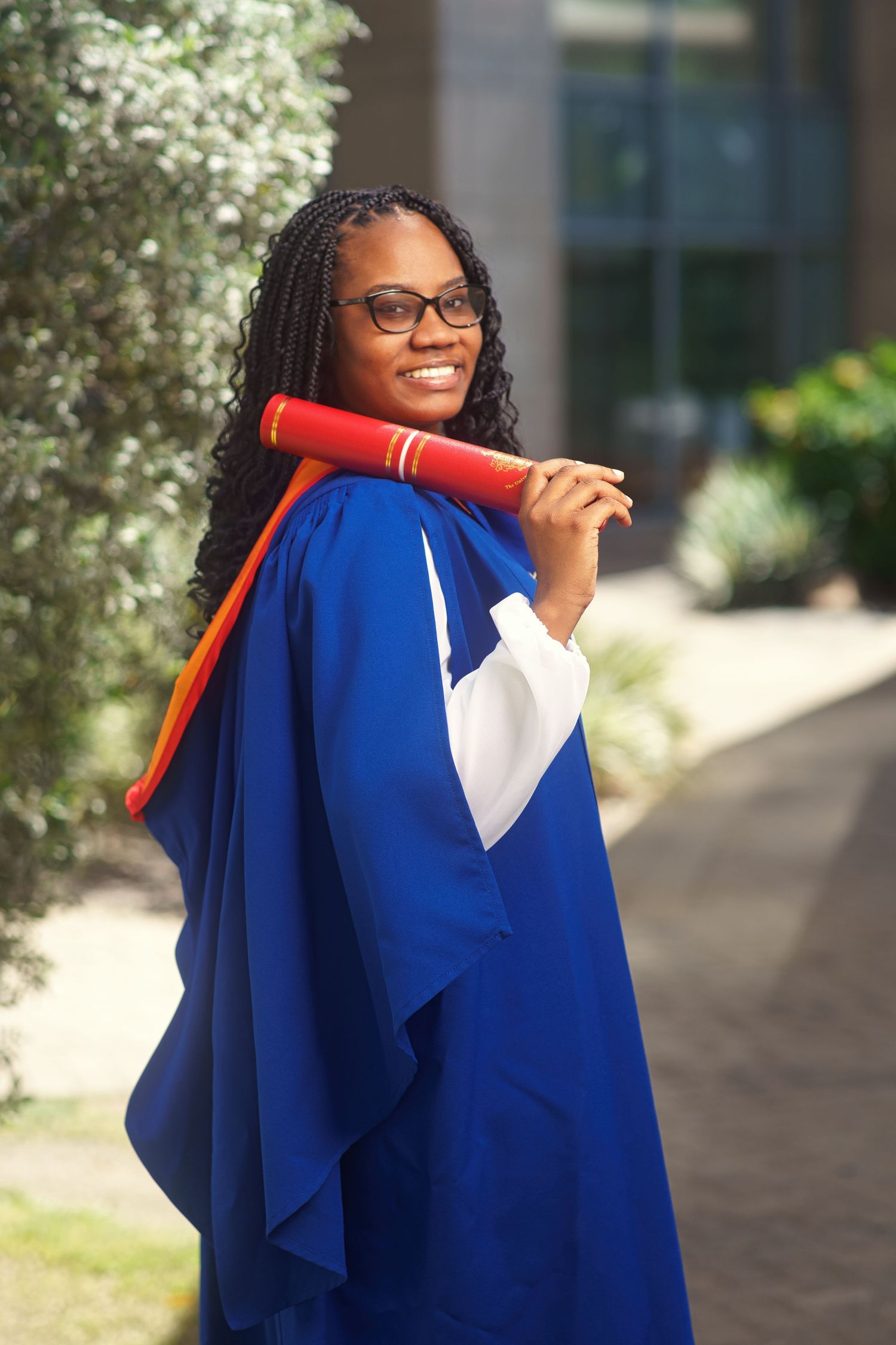 Graduate in blue academic regalia holds red diploma holder while smiling outdoors on campus pathway.