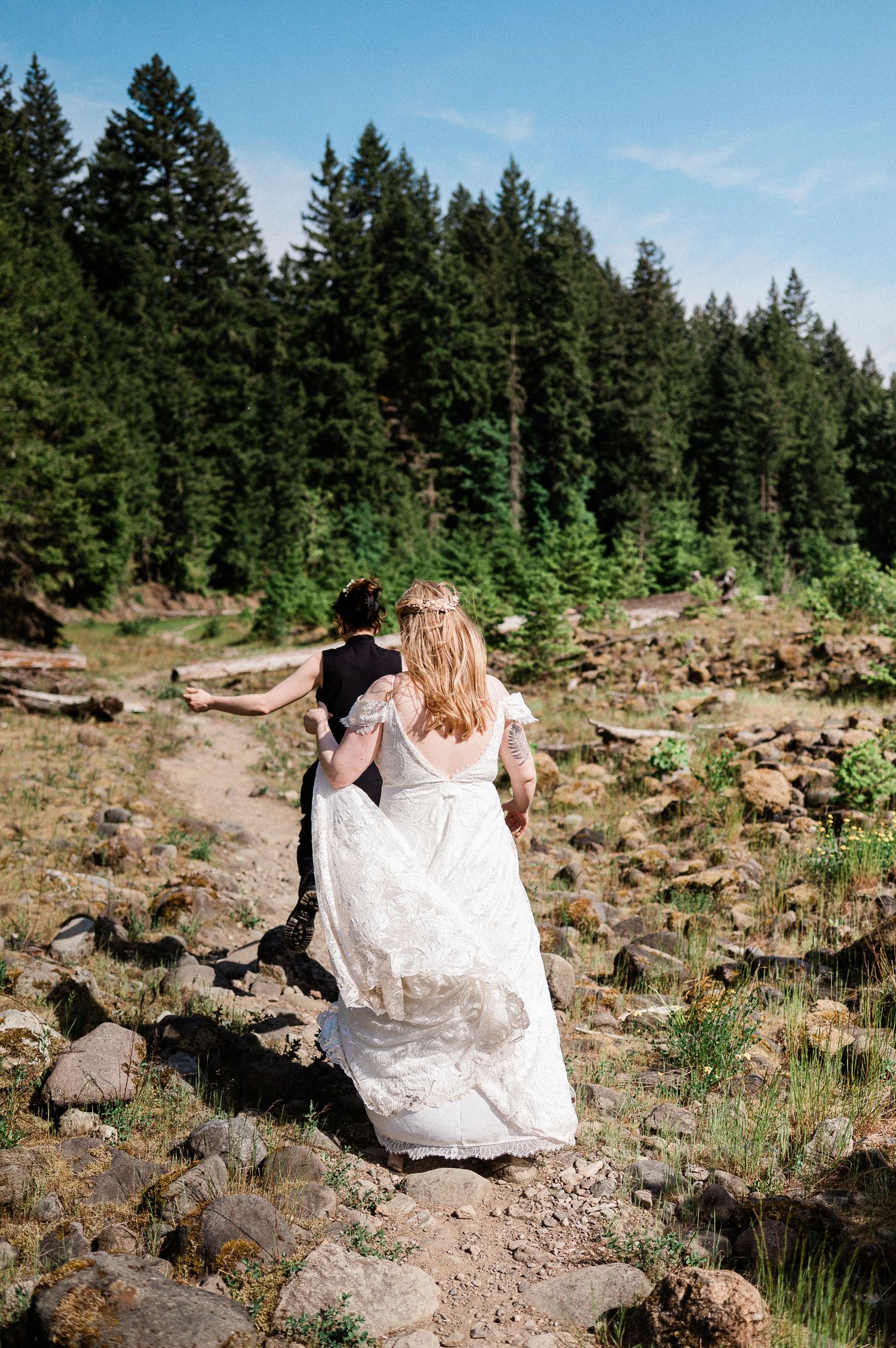 Two people hiking on a rocky path surrounded by evergreen trees on a sunny day.
