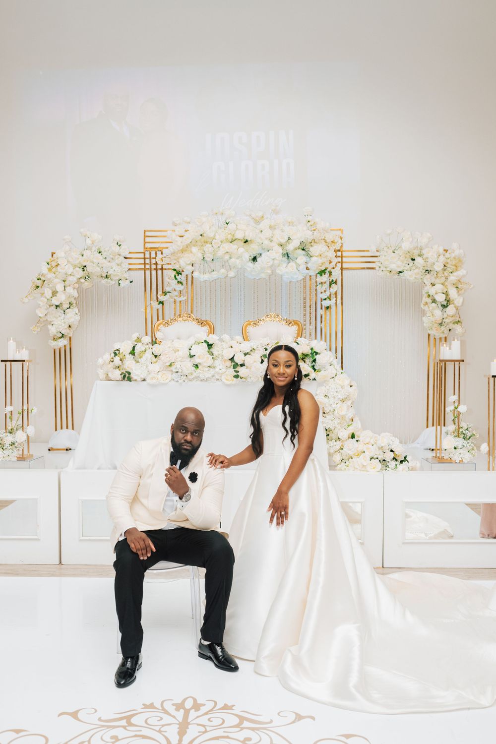 A luxurious wedding setup with white floral arch, gold accents and elegant decor against a light backdrop.