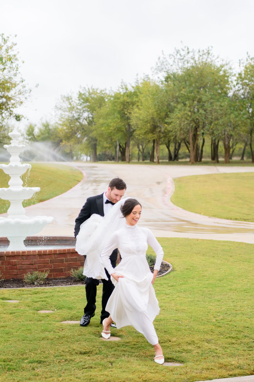 A couple in wedding attire playfully runs on a grassy lawn near a fountain on their special day.