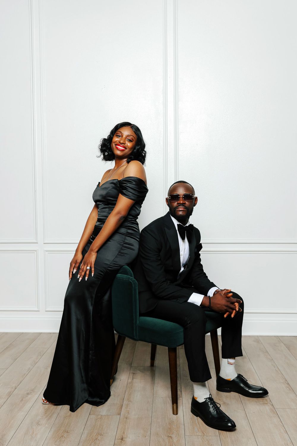 Elegant couple in formal black attire pose together against white wall, with emerald velvet chair accent.