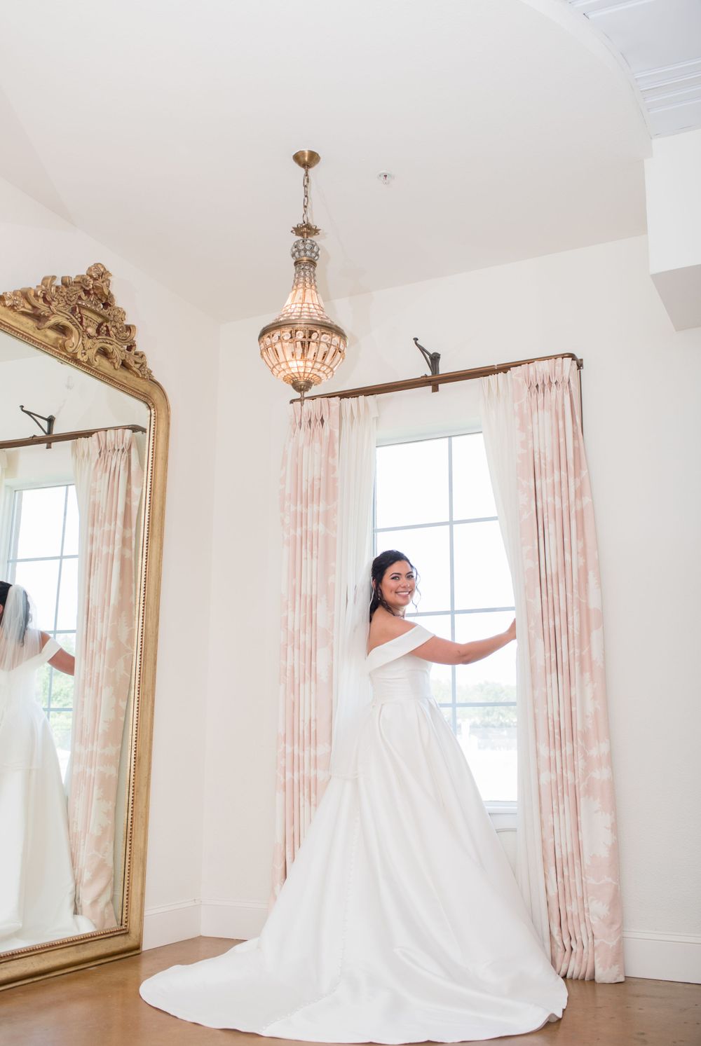 An elegant bridal portrait in a bright room with an ornate gold mirror, crystal chandelier and soft pink curtains.