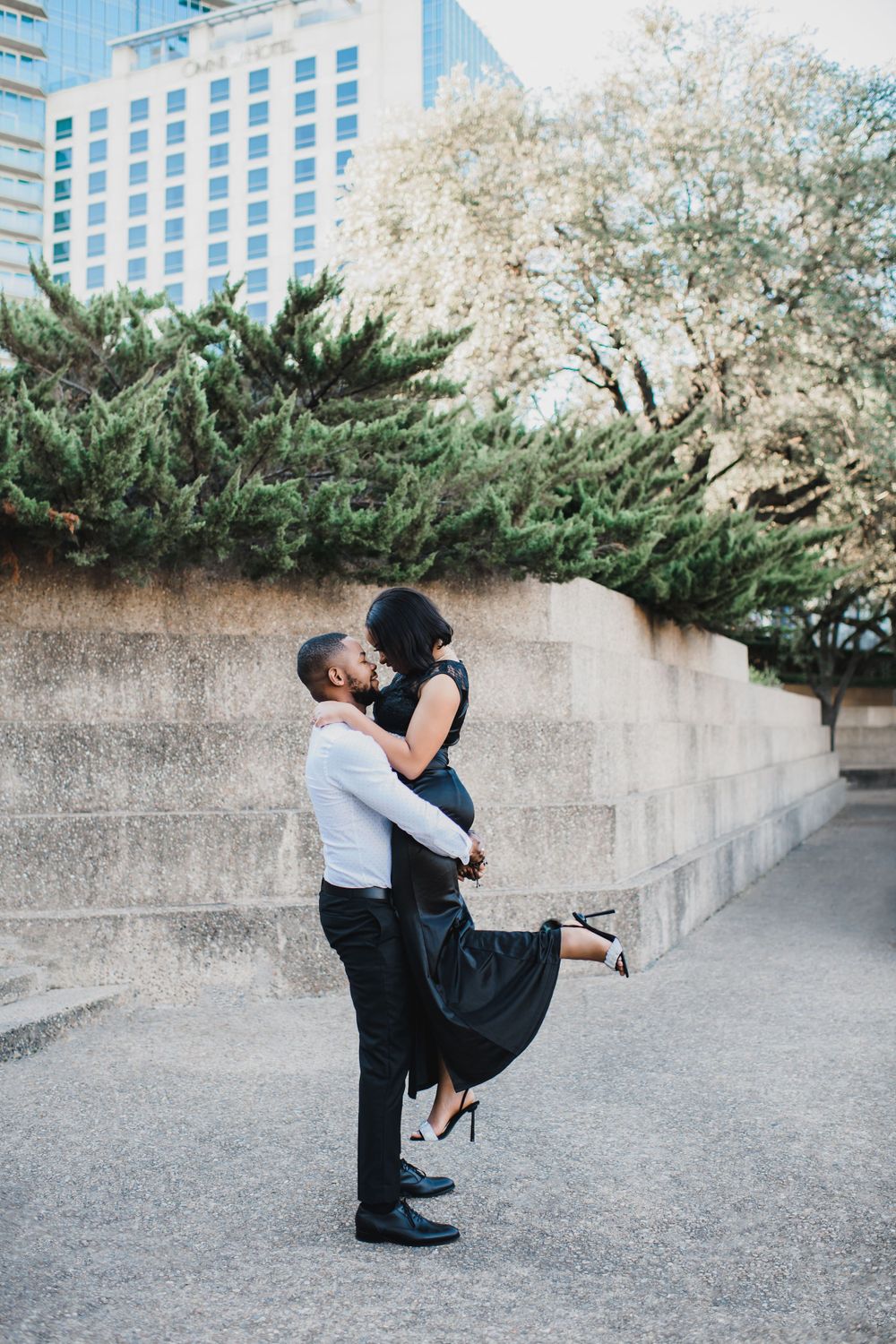 A romantic couple embraces in a lifting pose against a modern urban backdrop with trees and a tall building.