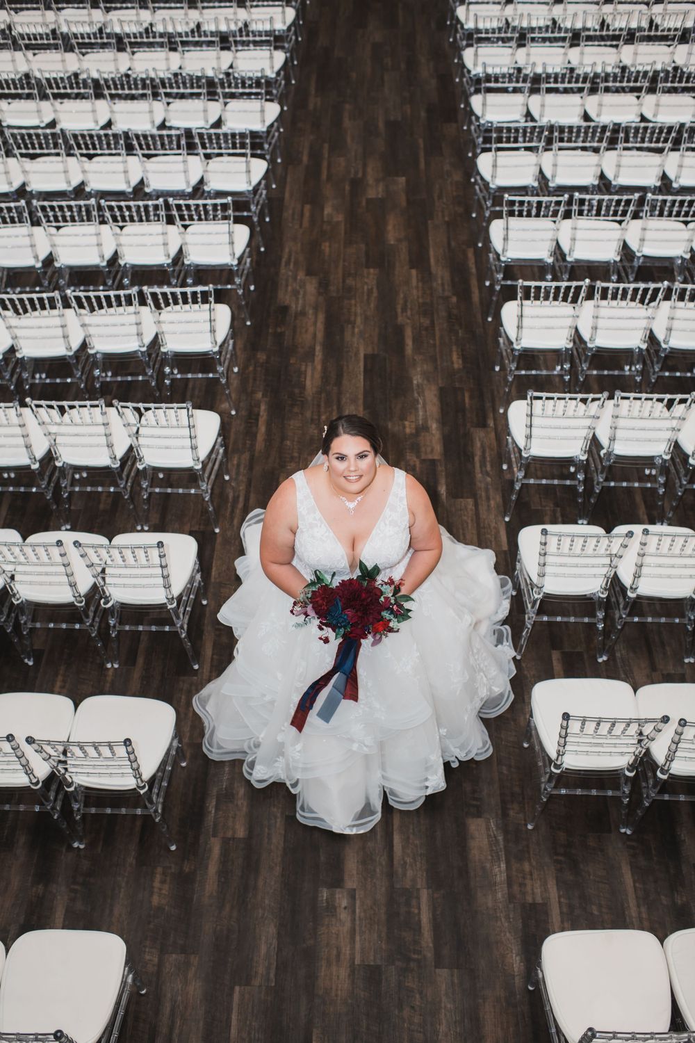 Aerial view of a person in a white wedding dress holding a bouquet while standing on a wooden floor among white chairs.