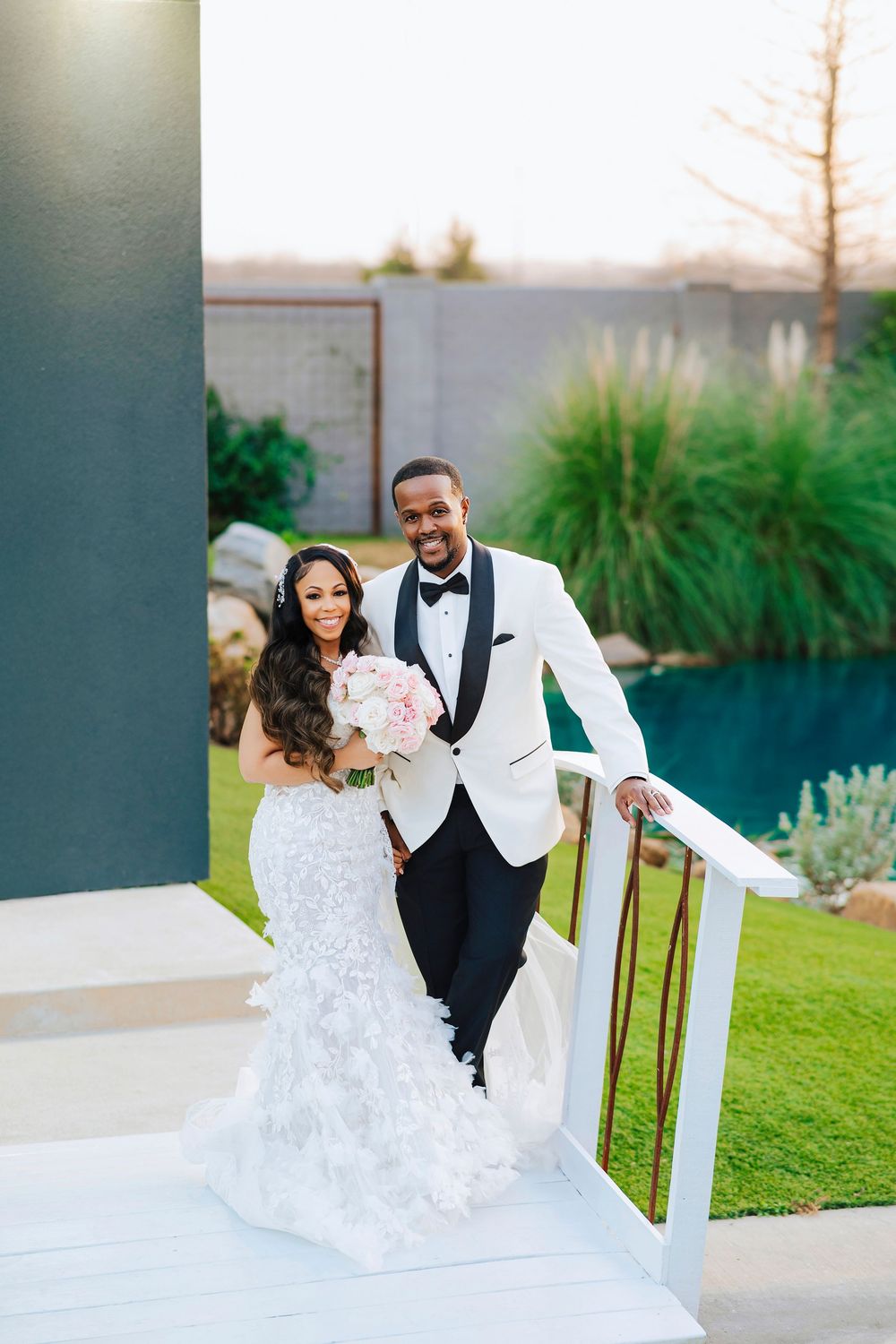 A wedding couple stands on outdoor steps in formal attire, with the bride in a lace gown and groom in white tuxedo jacket.
