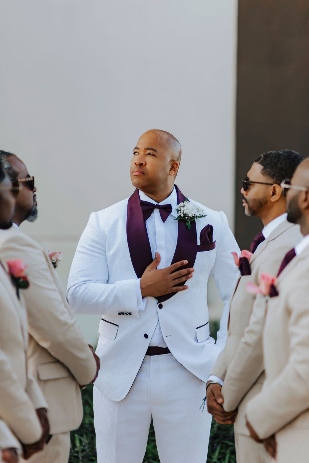 Wedding party members wear white suits with burgundy accents and stand together during outdoor celebration.
