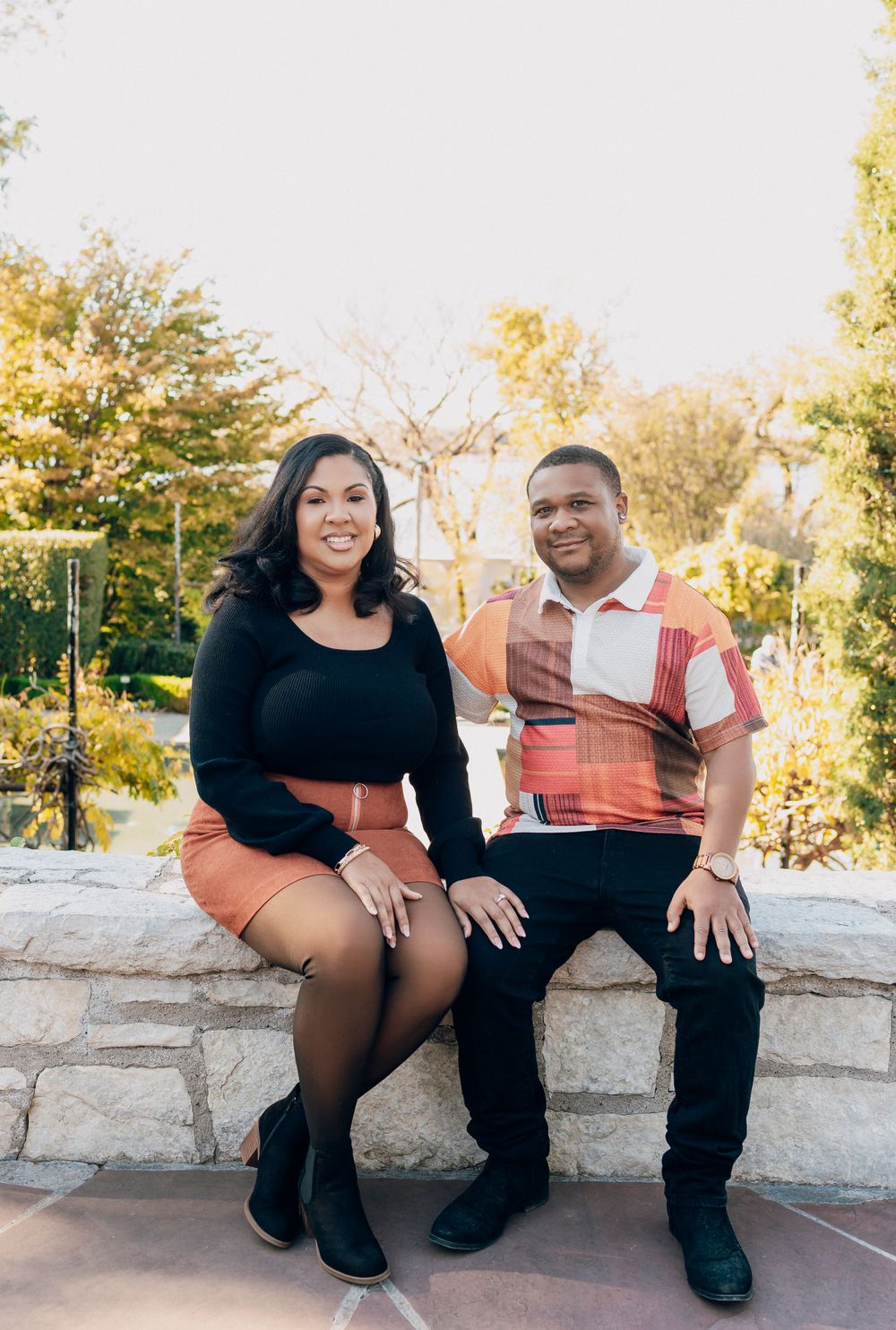 A couple sits together on a stone wall outdoors during sunset in a park setting with autumn trees in the background.