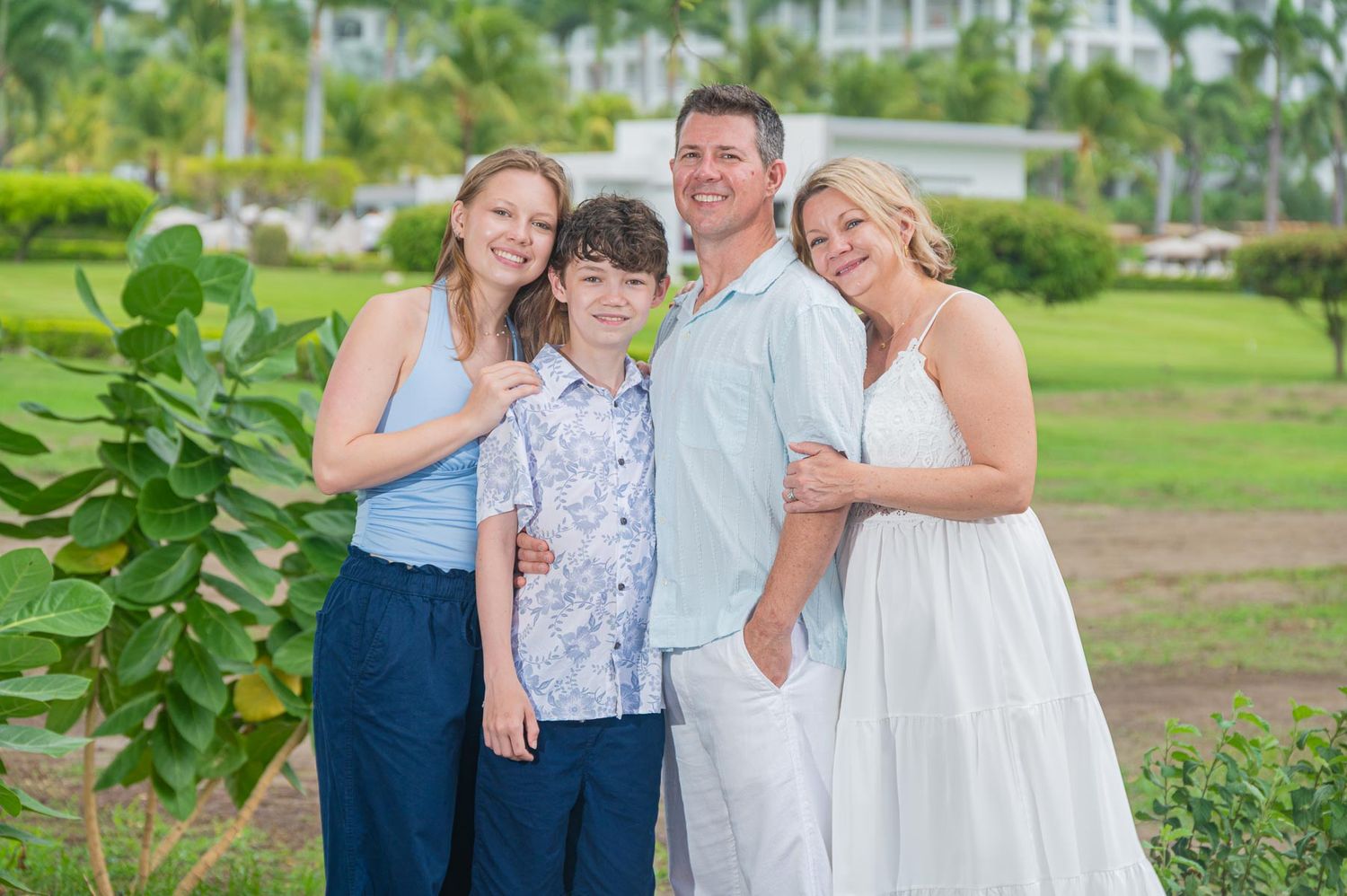 Family photoshoot of four smiling outdoors in tropical setting, wearing light summer clothing on a sunny day, at Riu