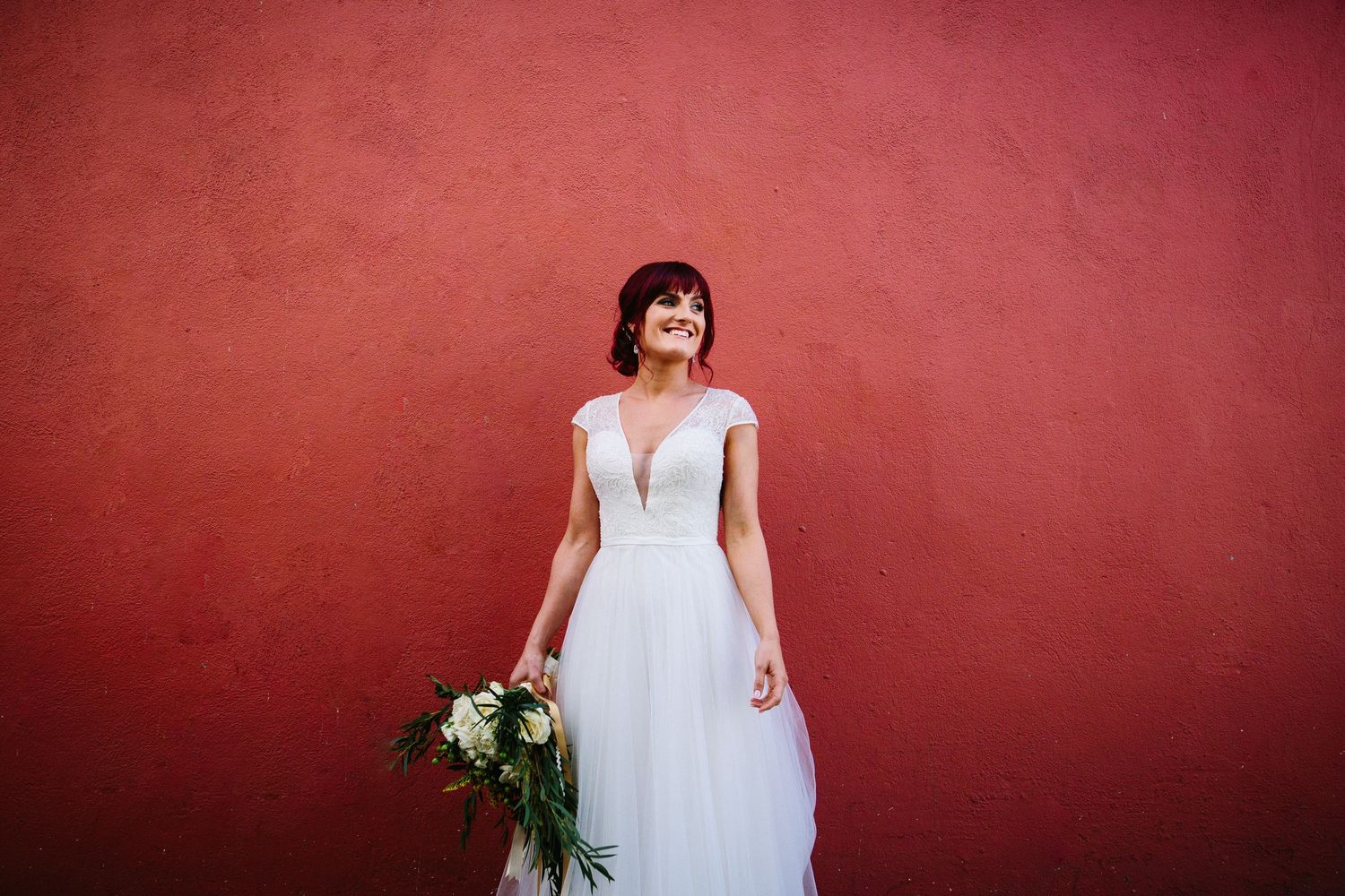 Bride in white gown holding bouquet smiles against vibrant red wall.