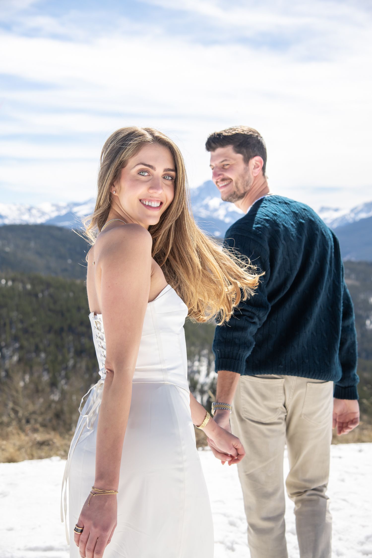 Couple holding hands in snowy mountain landscape, woman in white dress smiling over shoulder for Vail Colorado engagement photos.