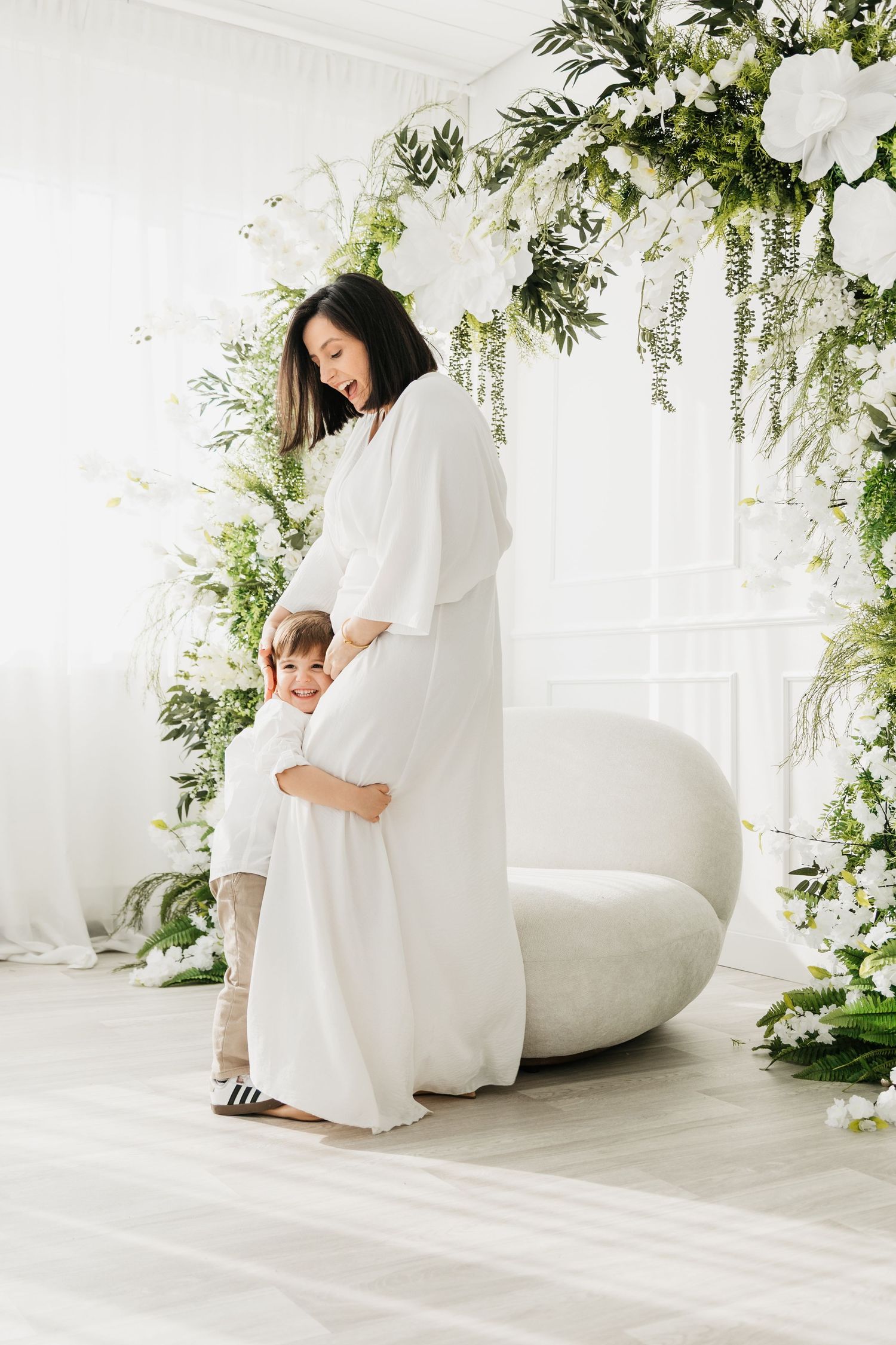 Pregnant woman in white dress hugging young child among white floral arch decor in bright studio.