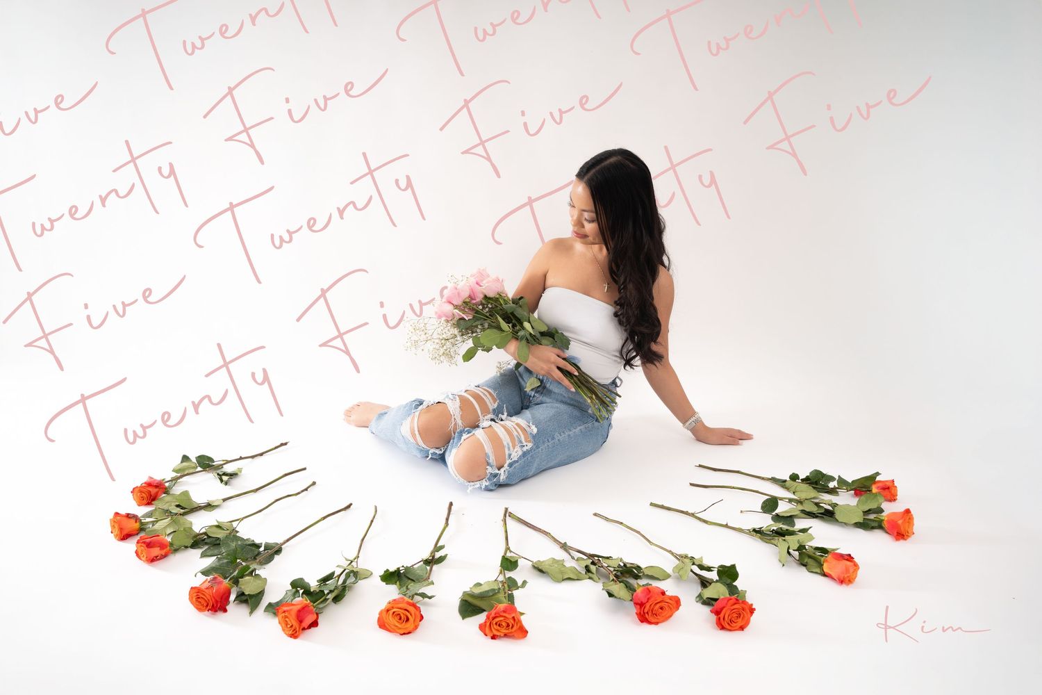A young woman poses sitting on a white backdrop in a studio setting surrounded by flowers with the words Twenty Five written across the imag