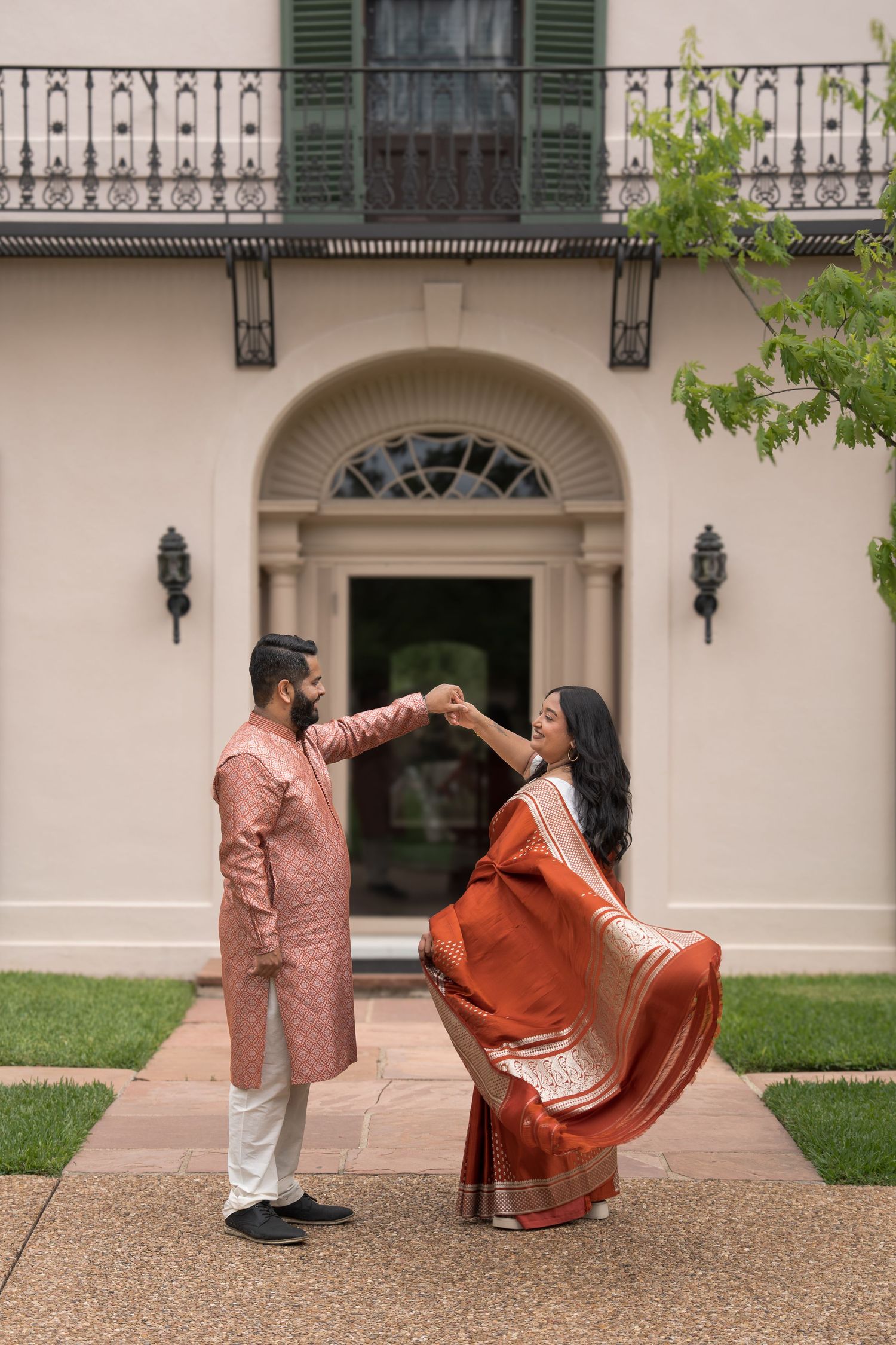 Indian couple in traditional attire dancing outside elegant mansion, man in pink kurta holding woman in rust saree.
