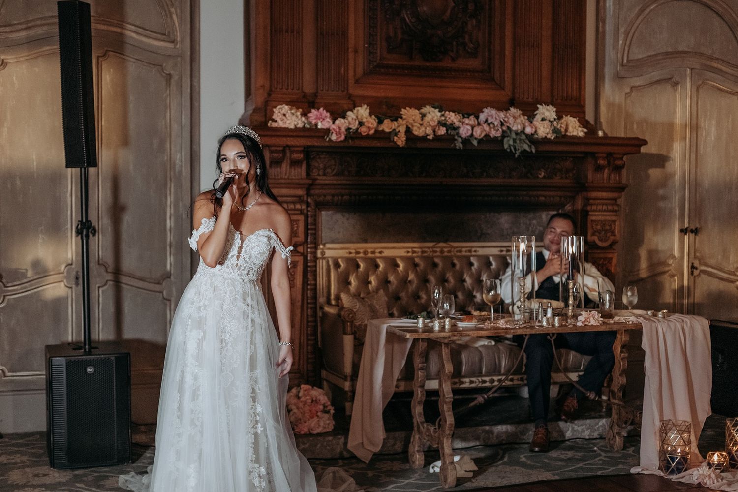 Bride in lace off-shoulder gown singing at wedding reception near ornate fireplace with floral decor at The Havens Country Club.