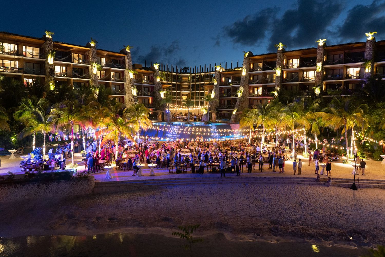 Outdoor evening incentive destination trip at a tropical resort with palm trees, colorful stage lights, and a large crowd on the beach.