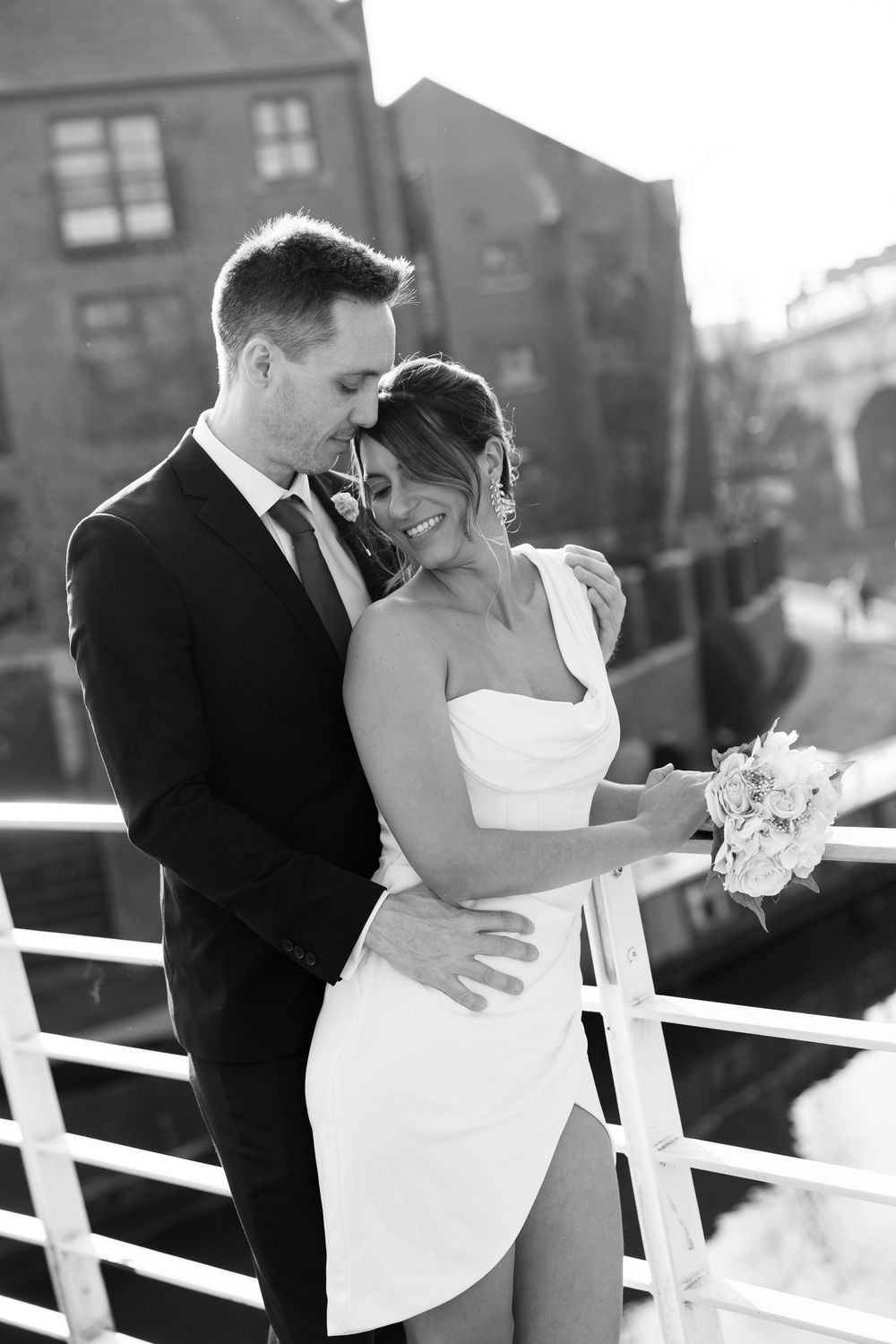 Groom embraces smiling bride holding bouquet on bridge in elegant black and white portrait.