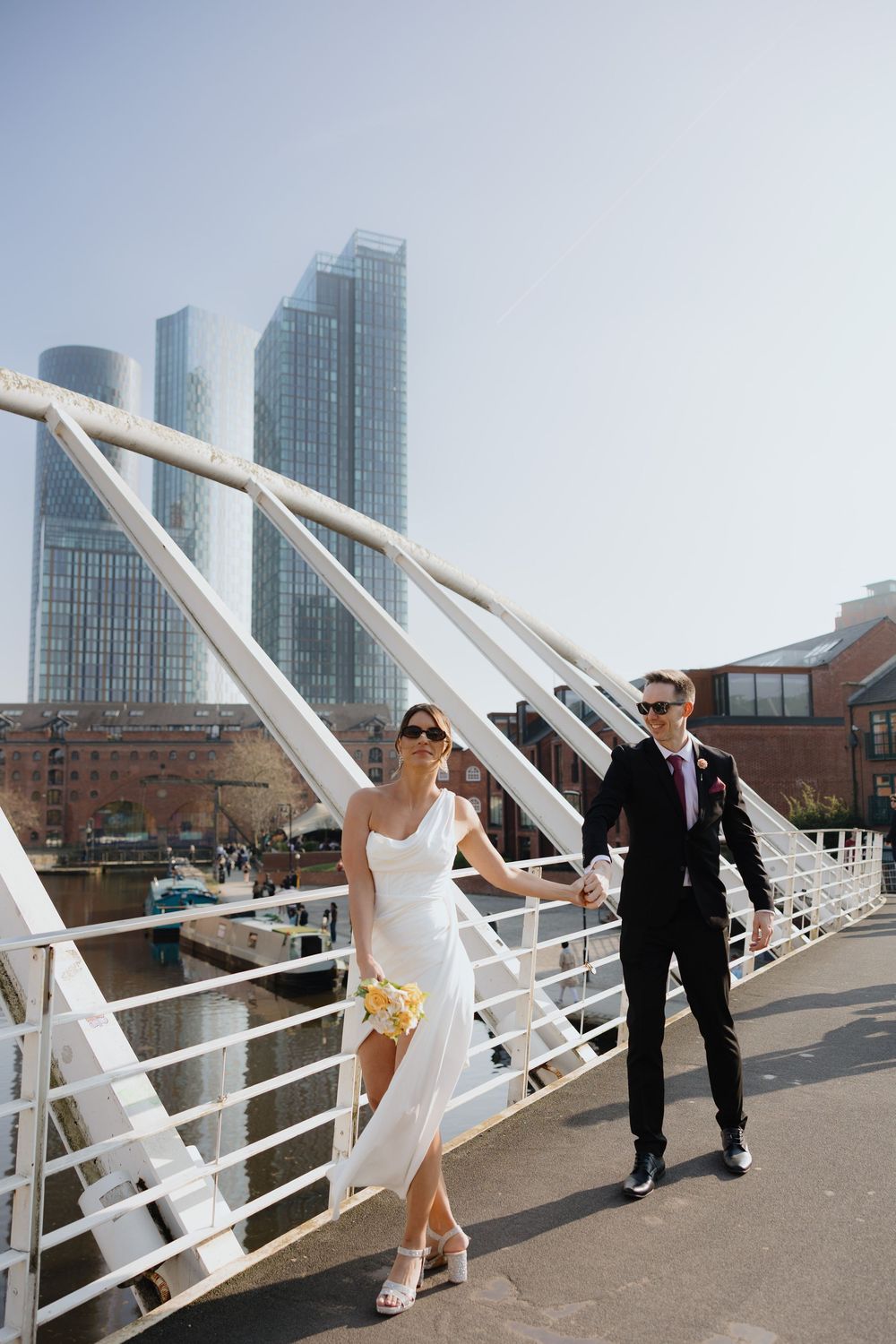 Bride and groom walking hand in hand on a modern white bridge with city skyscrapers in the background.