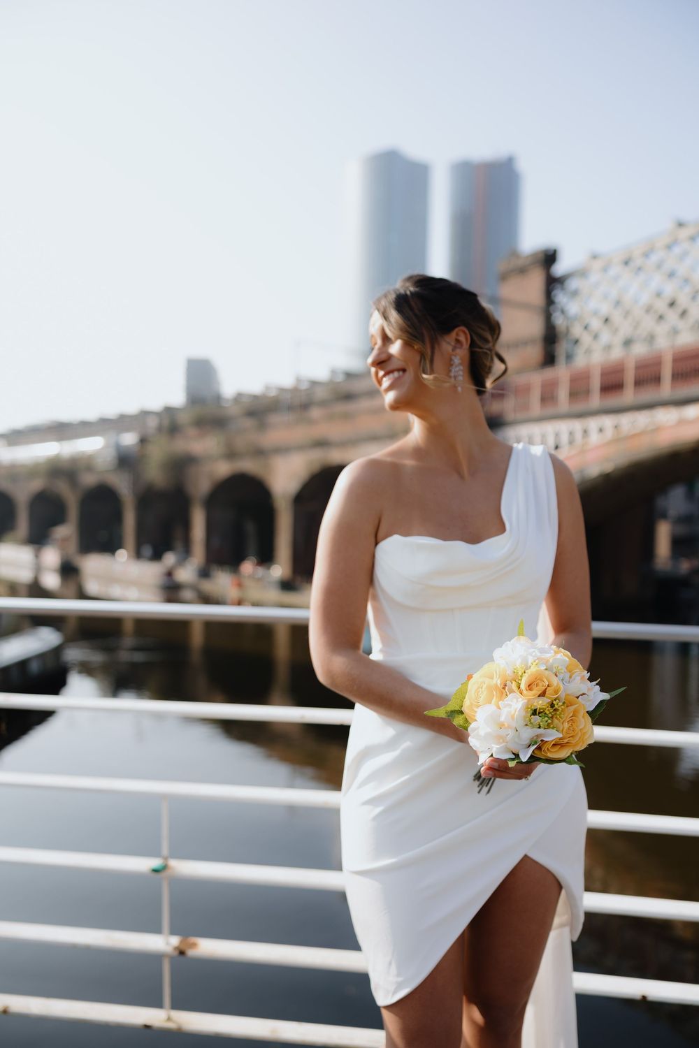 Smiling bride in white one-shoulder mini dress holding yellow bouquet on bridge with city skyline.