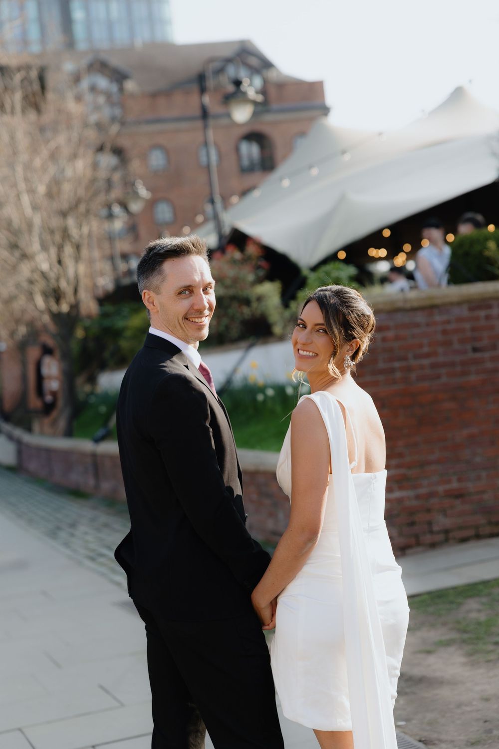 Smiling bride and groom holding hands and looking back at camera outside a wedding venue.