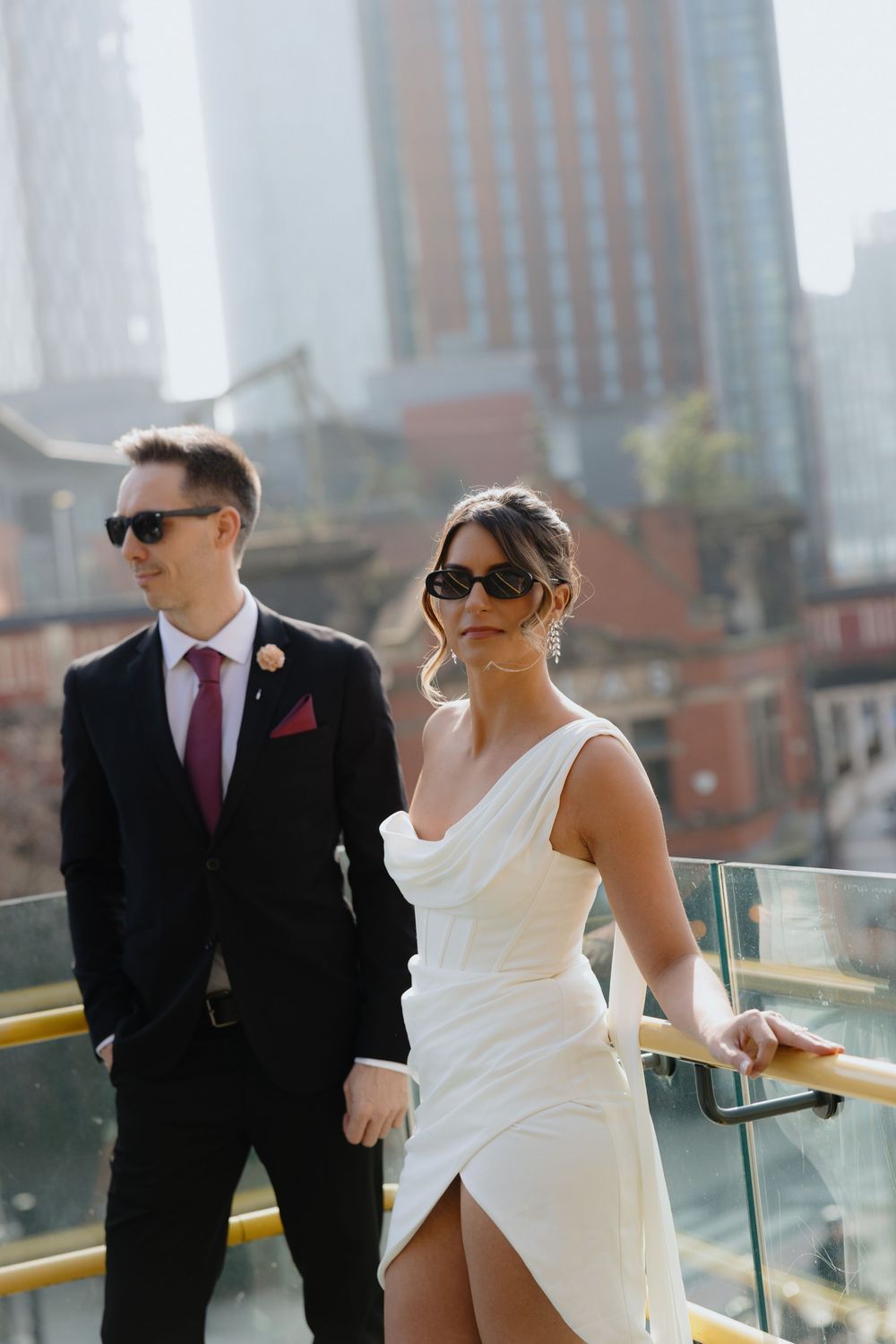 Stylish bride in white gown and groom in black suit wearing sunglasses on a rooftop city terrace.