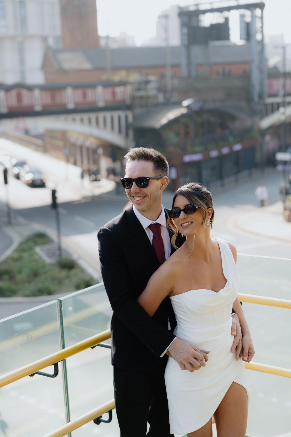 Smiling couple in wedding attire and sunglasses pose on a rooftop overlooking a city street and bridge.