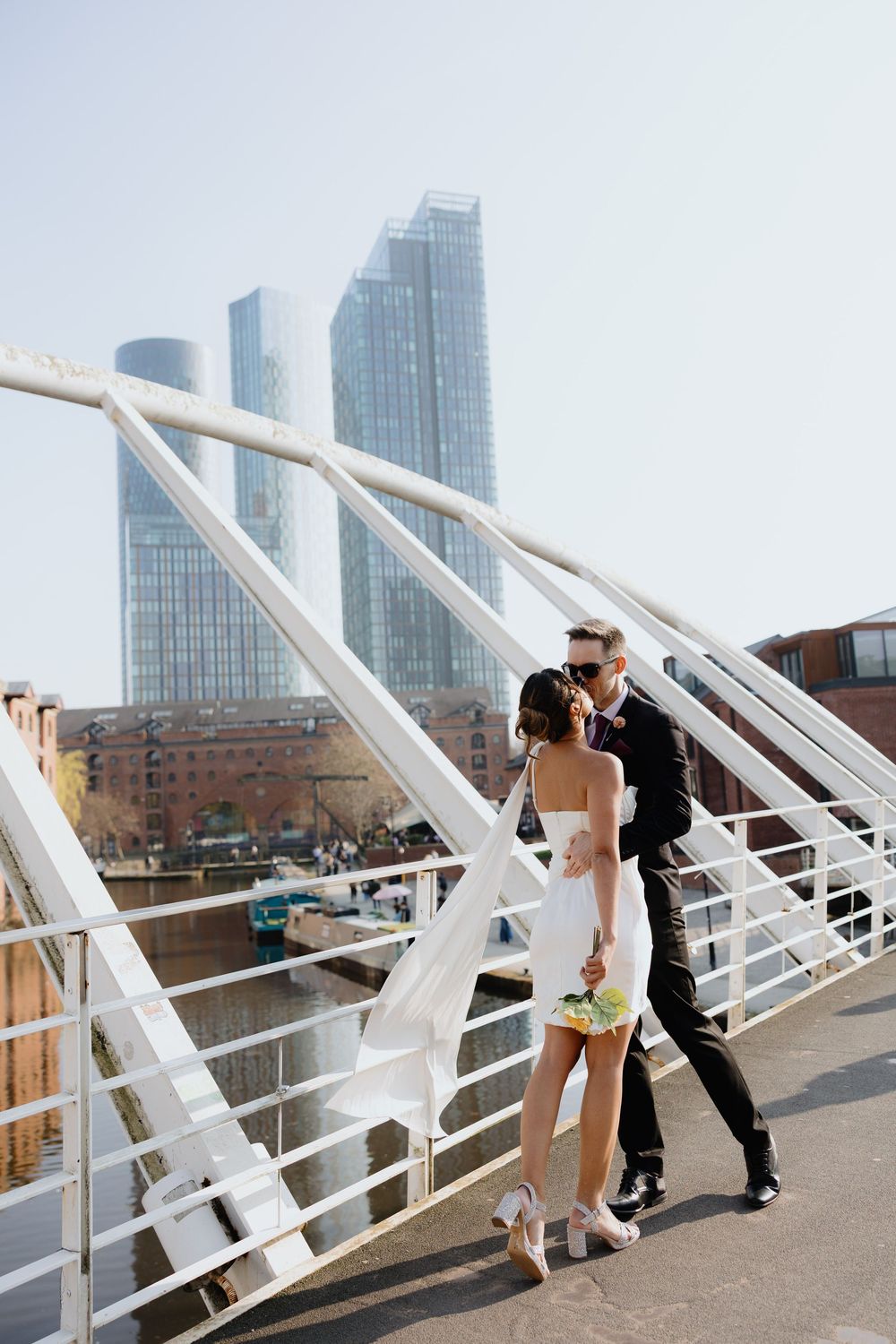 Wedding couple walking on white footbridge with modern city skyscrapers in background on sunny day.