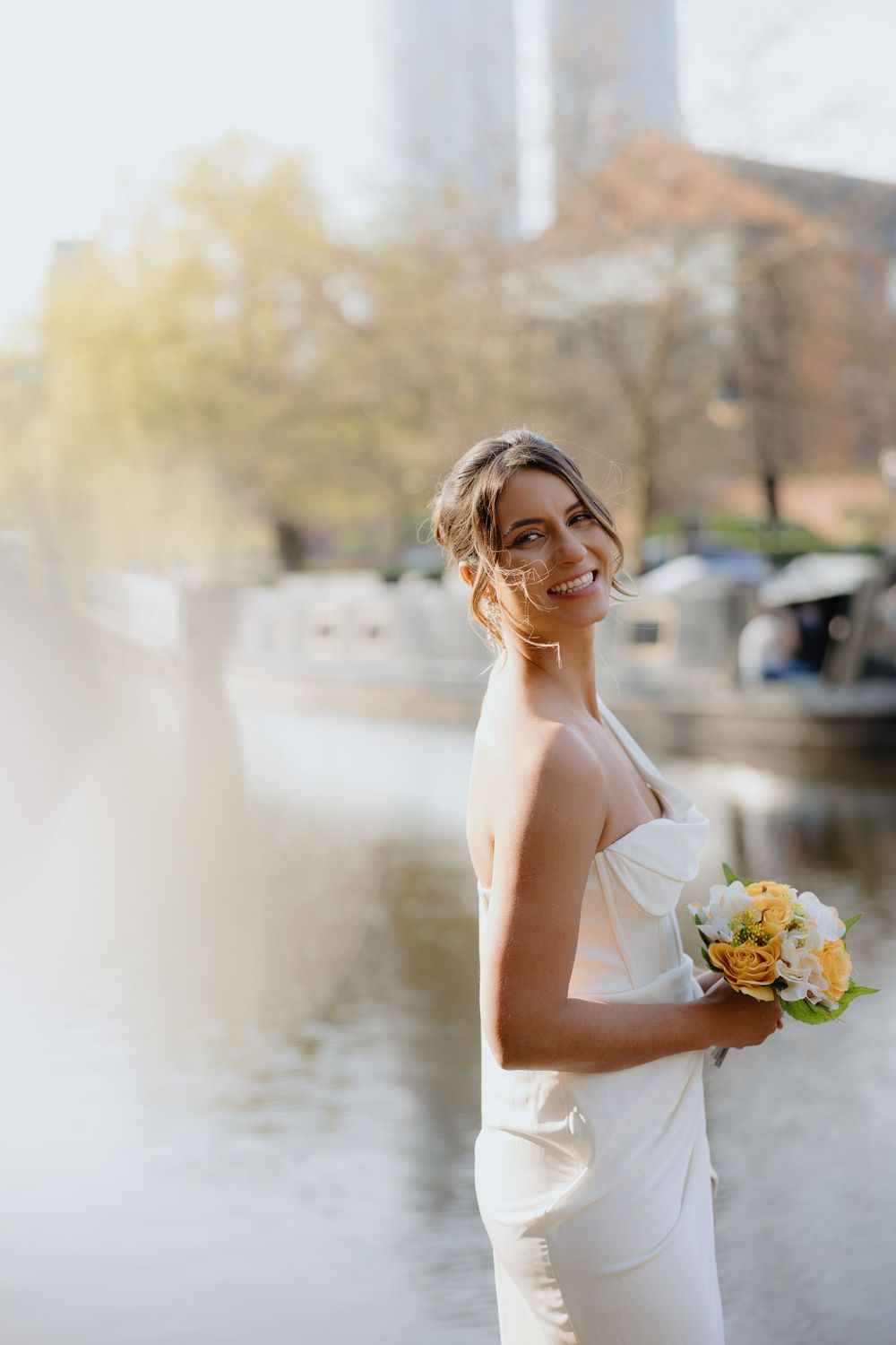 Smiling bride in white gown holds yellow bouquet beside urban waterway in golden sunlight.