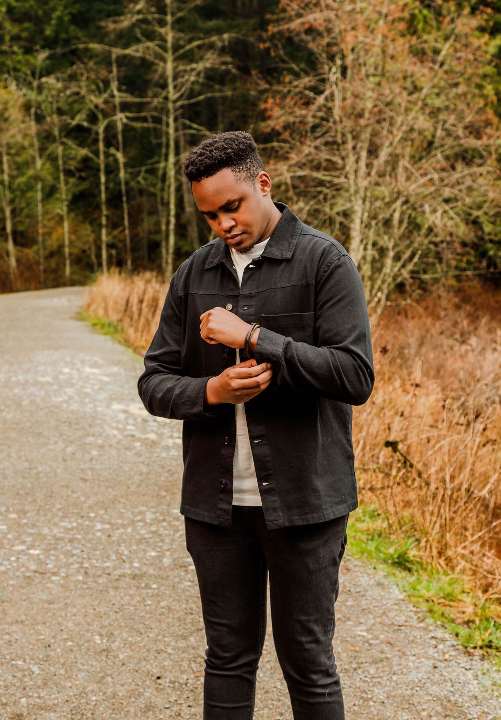 Young man in dark overshirt adjusts sleeve while standing on gravel path surrounded by autumn foliage.