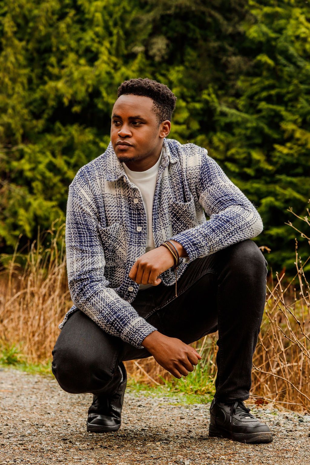 Young man in plaid shacket crouches on gravel path with evergreen trees in background during outdoor photoshoot.