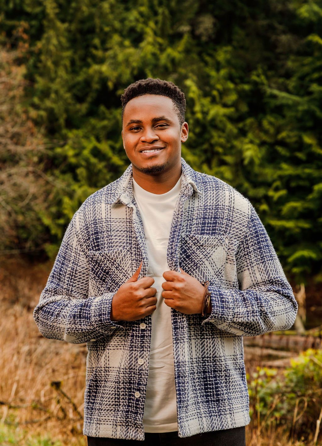 Smiling young man in blue plaid shacket poses outdoors with lush green evergreen trees in the background.