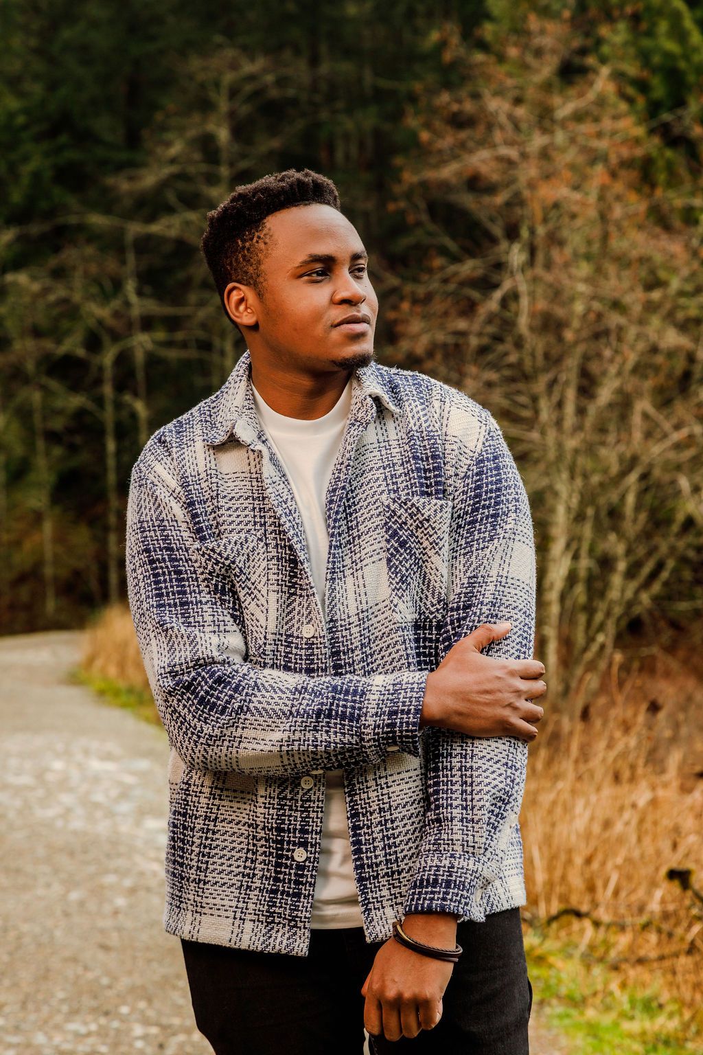 Young Black man in a blue plaid shacket stands on a forest path with arms crossed.
