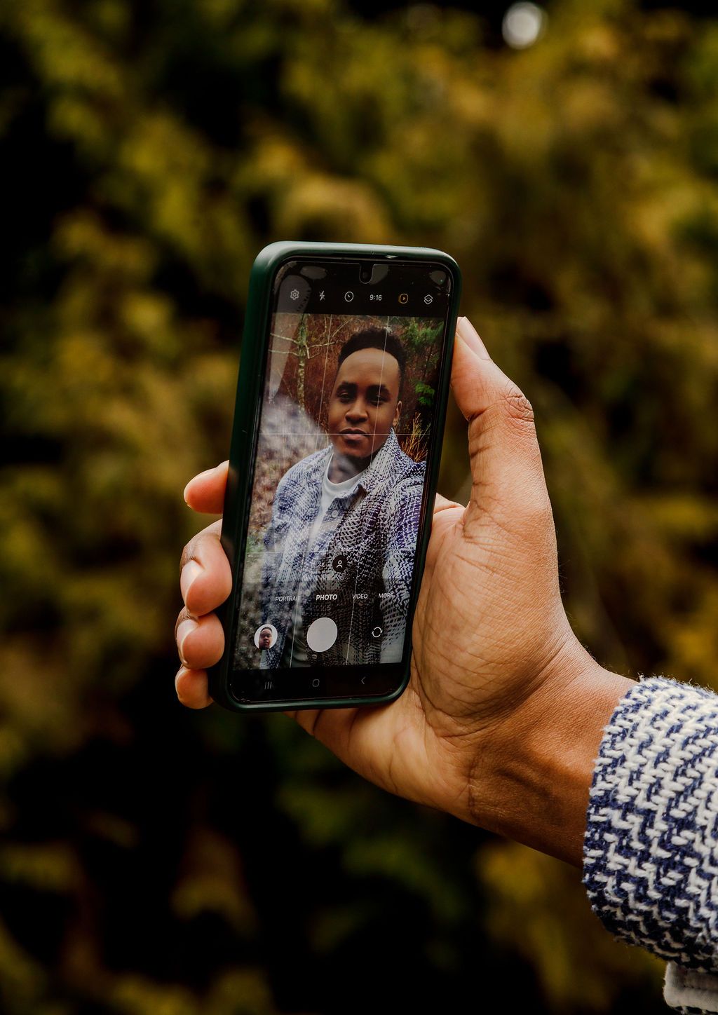 Hand holding a smartphone photographing a young Black man in a plaid jacket outdoors.