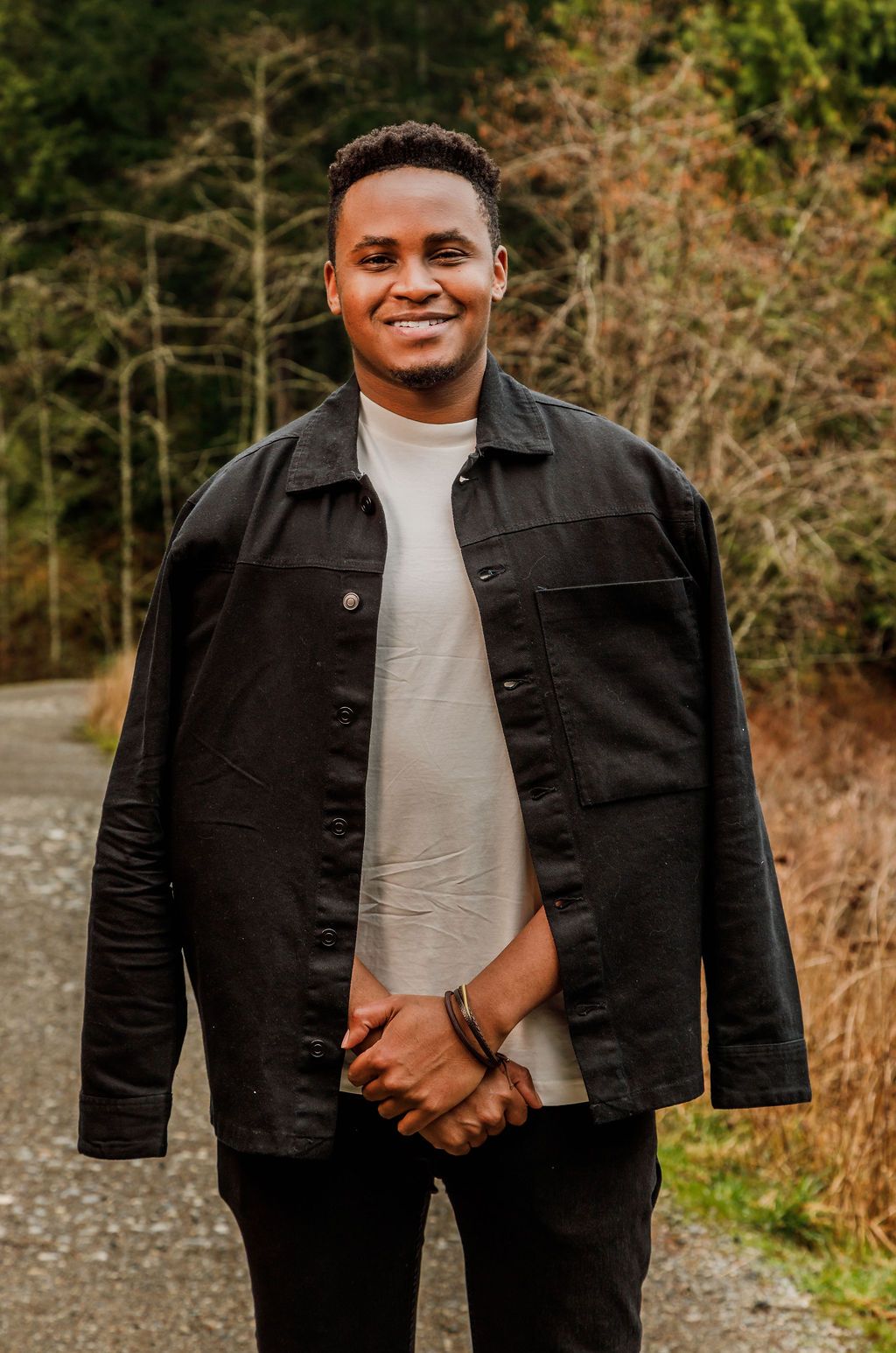 Smiling young Black man in a black jacket and cream turtleneck poses on a gravel path.