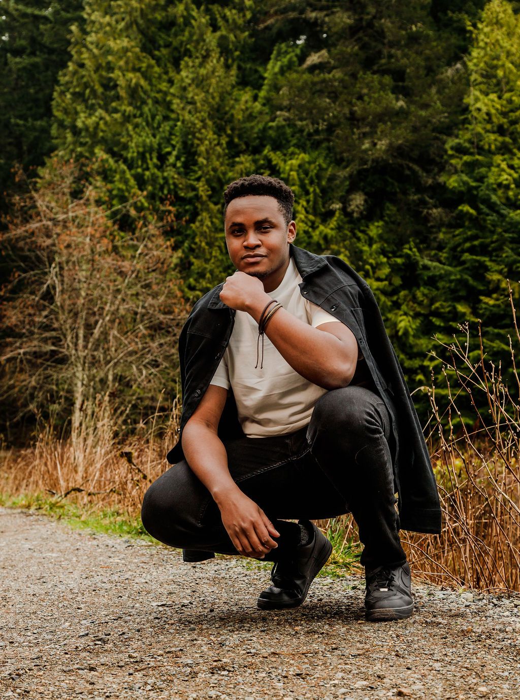 Young Black man in a black jacket crouches confidently on a gravel path surrounded by trees.