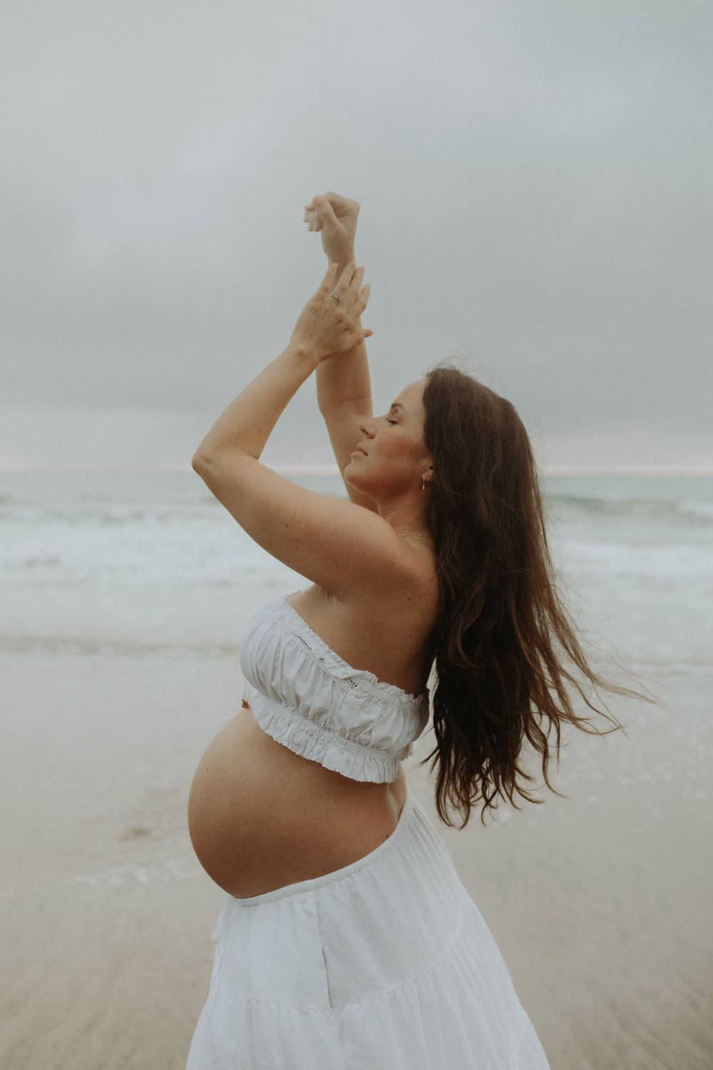 Pregnant woman in white crop top and skirt posing gracefully on a beach with arms raised, long dark hair blowing in wind.