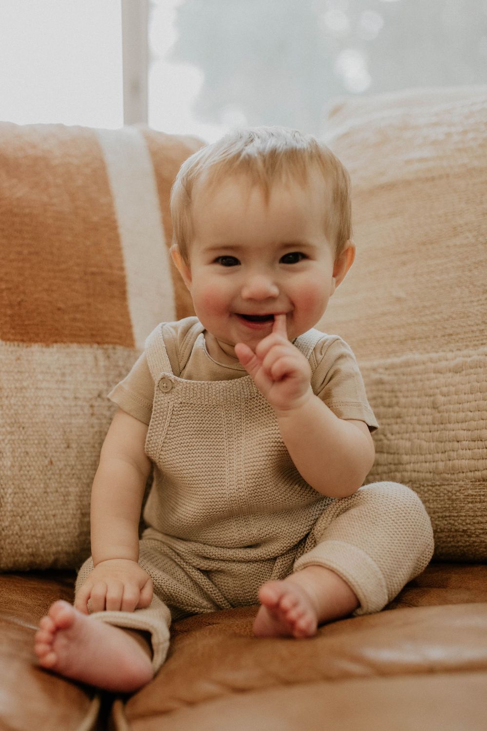 Smiling baby in beige knit romper sitting on leather sofa with neutral pillows, looking happy and playful.