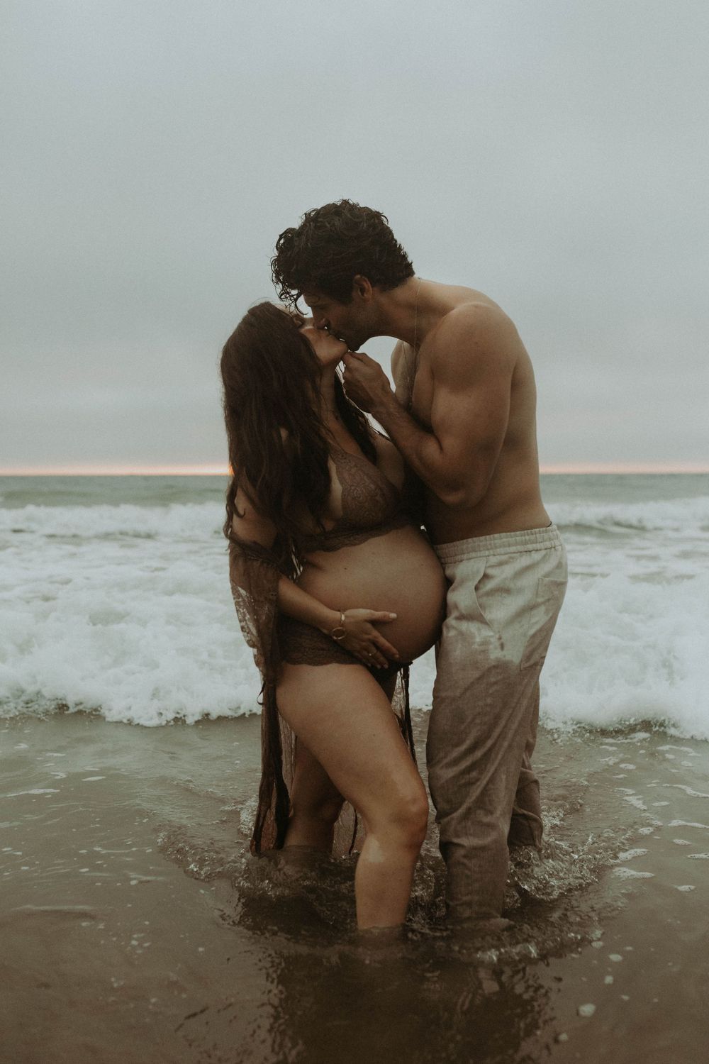 Pregnant couple sharing a kiss while standing in ocean waves on a cloudy beach day.