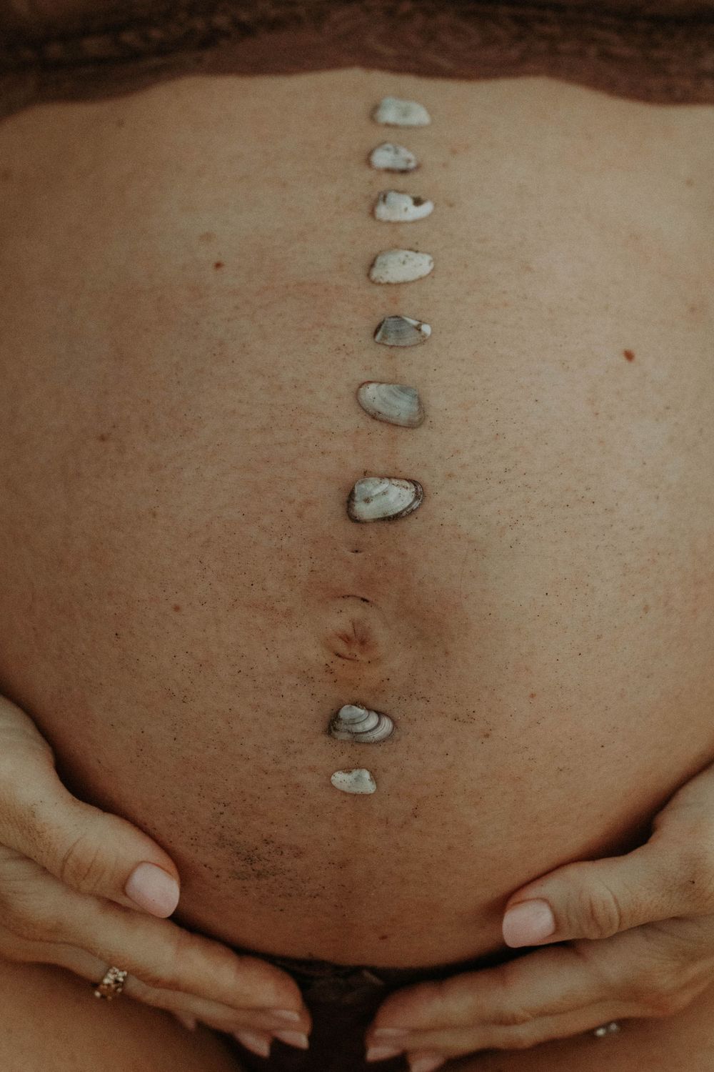 Pregnant woman's bare belly with small stones arranged vertically, hands gently cradling the bump.
