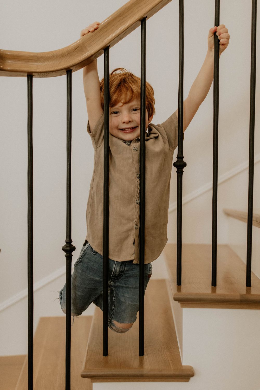 Joyful young child in tan shirt and jeans playing and hanging from wooden staircase with black metal balusters.