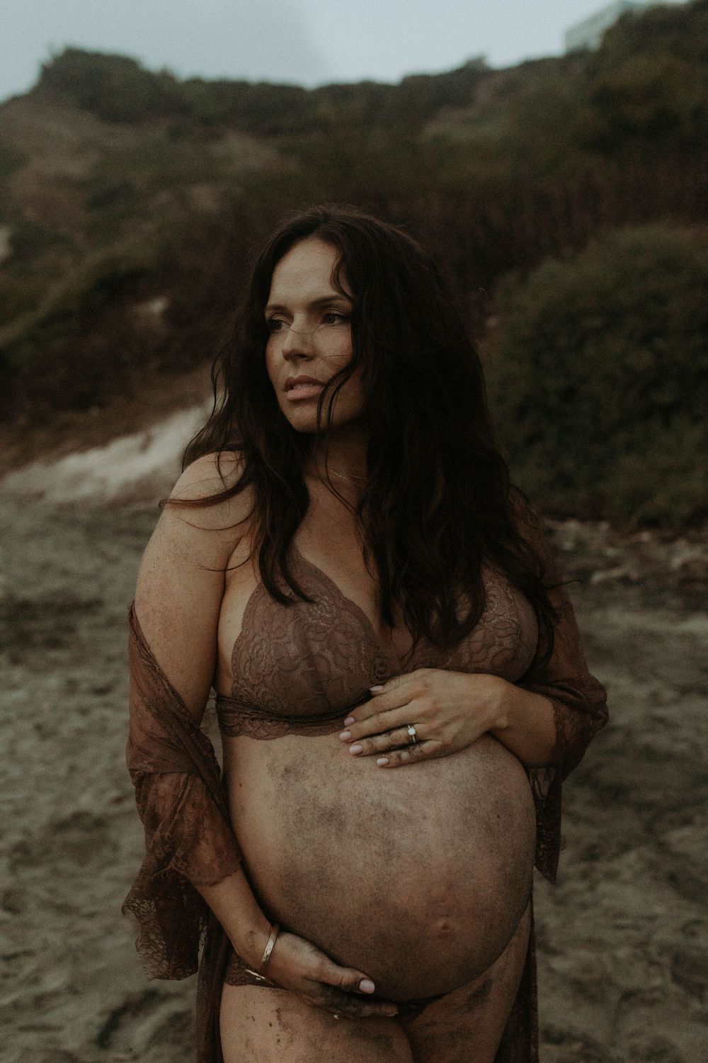 Pregnant woman in brown lace lingerie cradling her baby bump on a beach with green hills behind her.