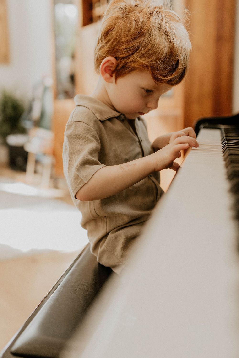Young toddler boy with red hair playing white piano keys in a warmly lit room wearing a beige polo shirt.