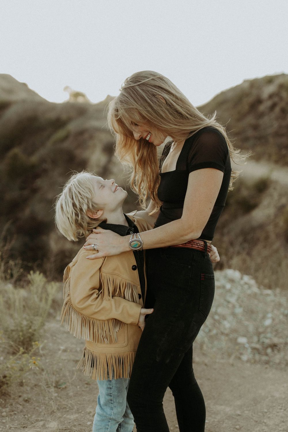 A tender outdoor moment between parent and child in nature, with the child wearing a fringed tan jacket and denim.